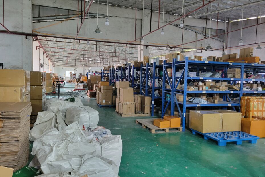 Warehouse interior with blue shelving units filled with cardboard boxes and pallets, showing goods consolidation from multiple suppliers for shipping to one central warehouse.