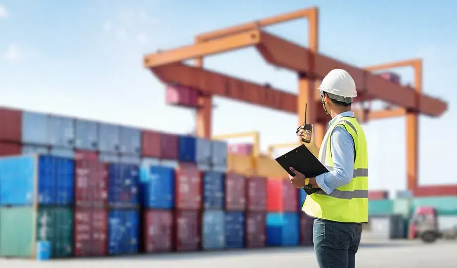 A logistics supervisor in a safety vest using a walkie-talkie and clipboard in a container yard, symbolizing the operational management of a Guangzhou warehouse shipping agent.