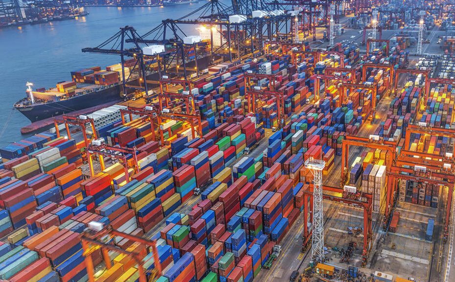 An aerial twilight shot of a densely packed container port with colorful containers, cranes, and a docked ship, representing the global logistics infrastructure behind DDP cost.